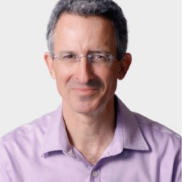 A middle-aged man with short, gray hair and glasses, wearing a light purple collared shirt, is smiling at the camera against a plain background.