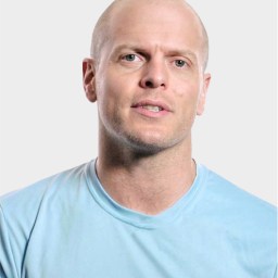 A man with a shaved head wearing a light blue t-shirt is facing the camera against a plain light background.