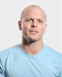 A man with a shaved head wearing a light blue t-shirt is facing the camera against a plain light background.