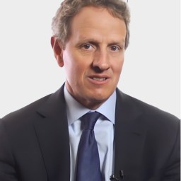 A man in a suit and tie is speaking, looking slightly to the side, with a neutral background.