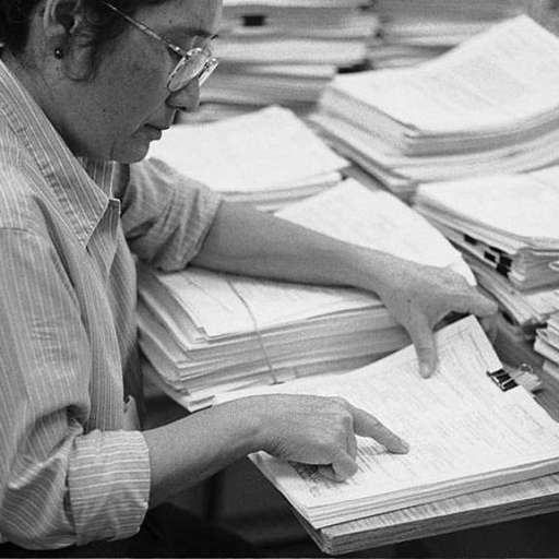A woman sits at a desk covered with tall stacks of papers, reviewing and pointing to documents as she conducts a purpose-driven peer review in a busy office setting.