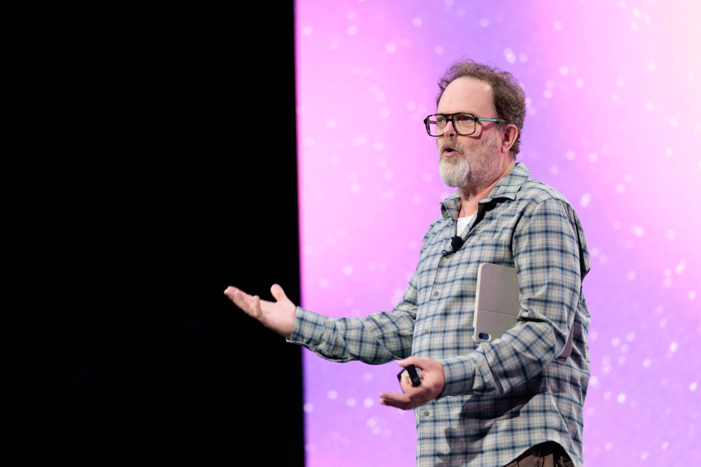 A man in a plaid shirt speaks on stage with a tablet under his arm, gesturing with his hands, in front of a purple backdrop.