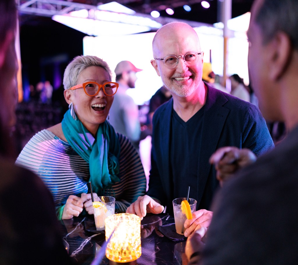 Two people stand at a table with drinks, smiling and conversing with others at a lively indoor event. A candle and another drink are on the table.