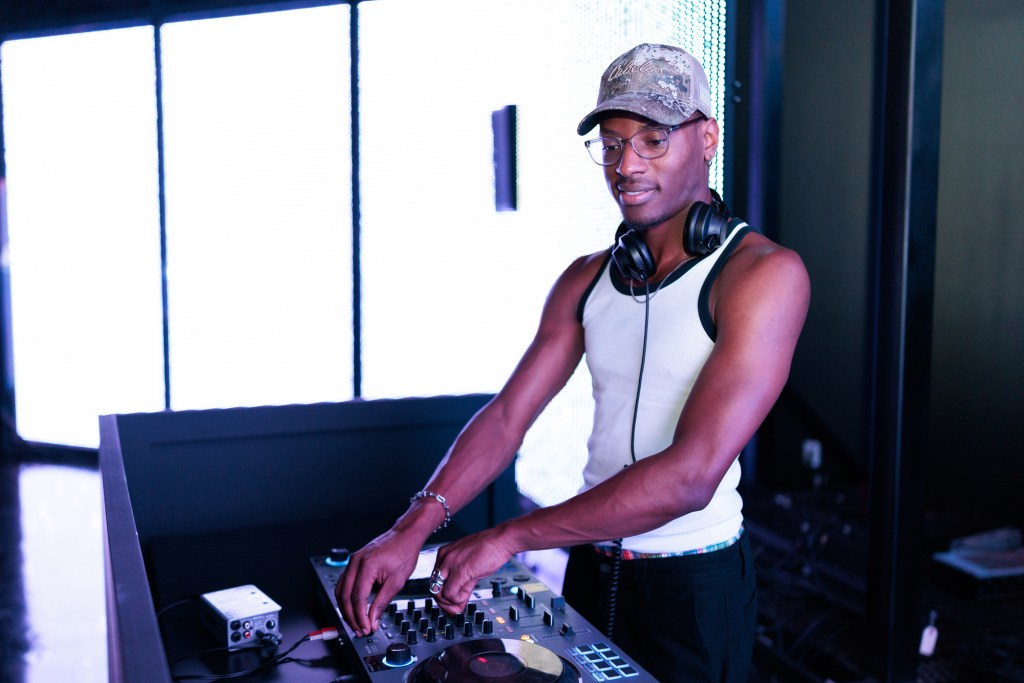 A DJ wearing a tank top, cap, and headphones stands at a mixing console, adjusting controls in a modern indoor setting.