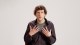 A person with curly brown hair wearing a black shirt and gray t-shirt sits against a plain white background, gesturing with both hands raised near their chest.