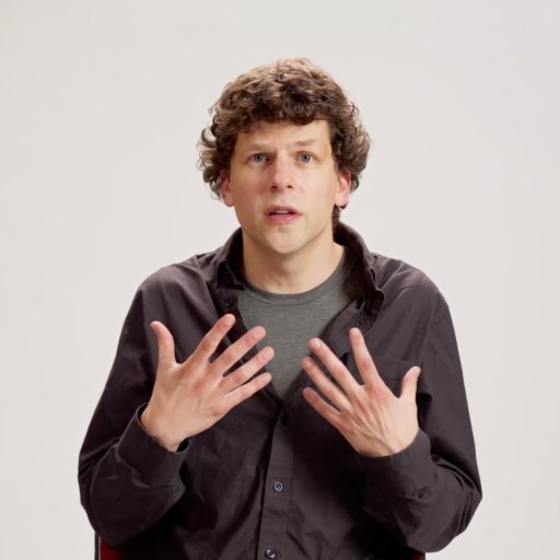 A person with curly brown hair wearing a black shirt and gray t-shirt sits against a plain white background, gesturing with both hands raised near their chest.