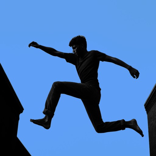 A person is leaping between two buildings against a clear blue sky.