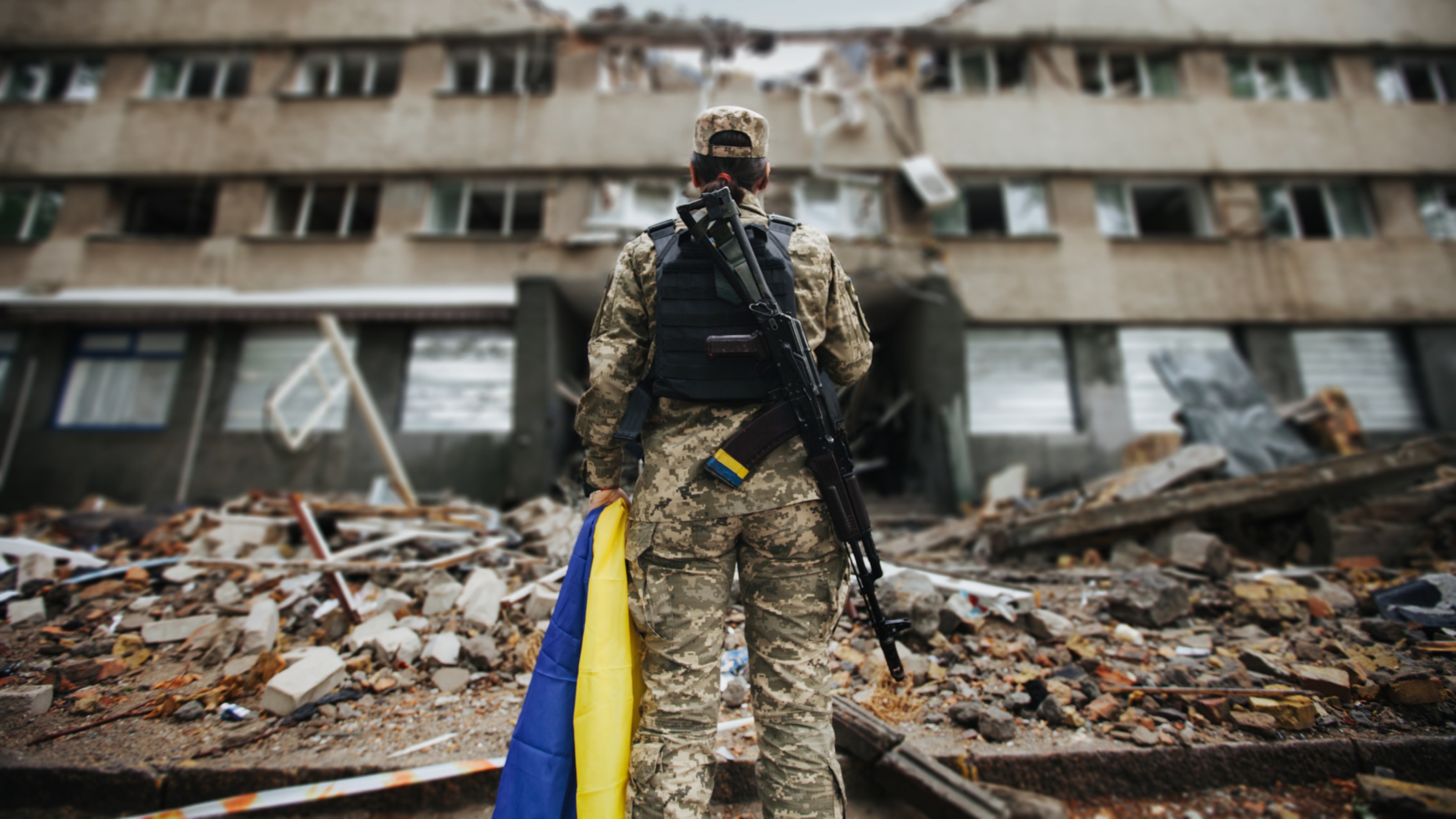 A soldier in camouflage uniform holding a Ukrainian flag stands amid rubble, facing a damaged building.