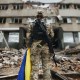 A soldier in camouflage uniform holding a Ukrainian flag stands amid rubble, facing a damaged building.