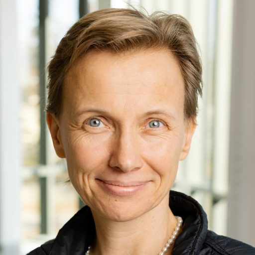 A woman with short light brown hair, wearing a black vest with a "sci foo 2019" patch and a pearl necklace, standing indoors in front of large windows.