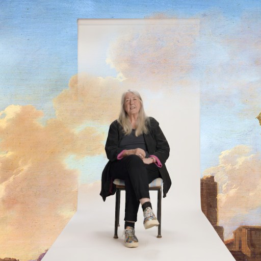 A person with long white hair sits smiling on a chair, set against a painted backdrop of classical architecture and a sky with clouds.