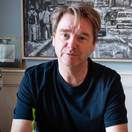 A man with light brown hair wearing a black t-shirt sits indoors in front of a black and white painting and a framed photo.