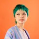 Person with short green hair and a pastel striped jacket looks slightly to the side against a solid orange background.