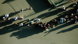 Aerial view of two groups of people waiting in line and walking on a paved surface, casting long shadows in the sunlight.