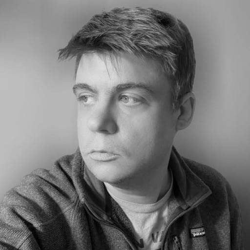 A man with short hair and a zip-up jacket looks to his left against a plain background in a black and white photo.