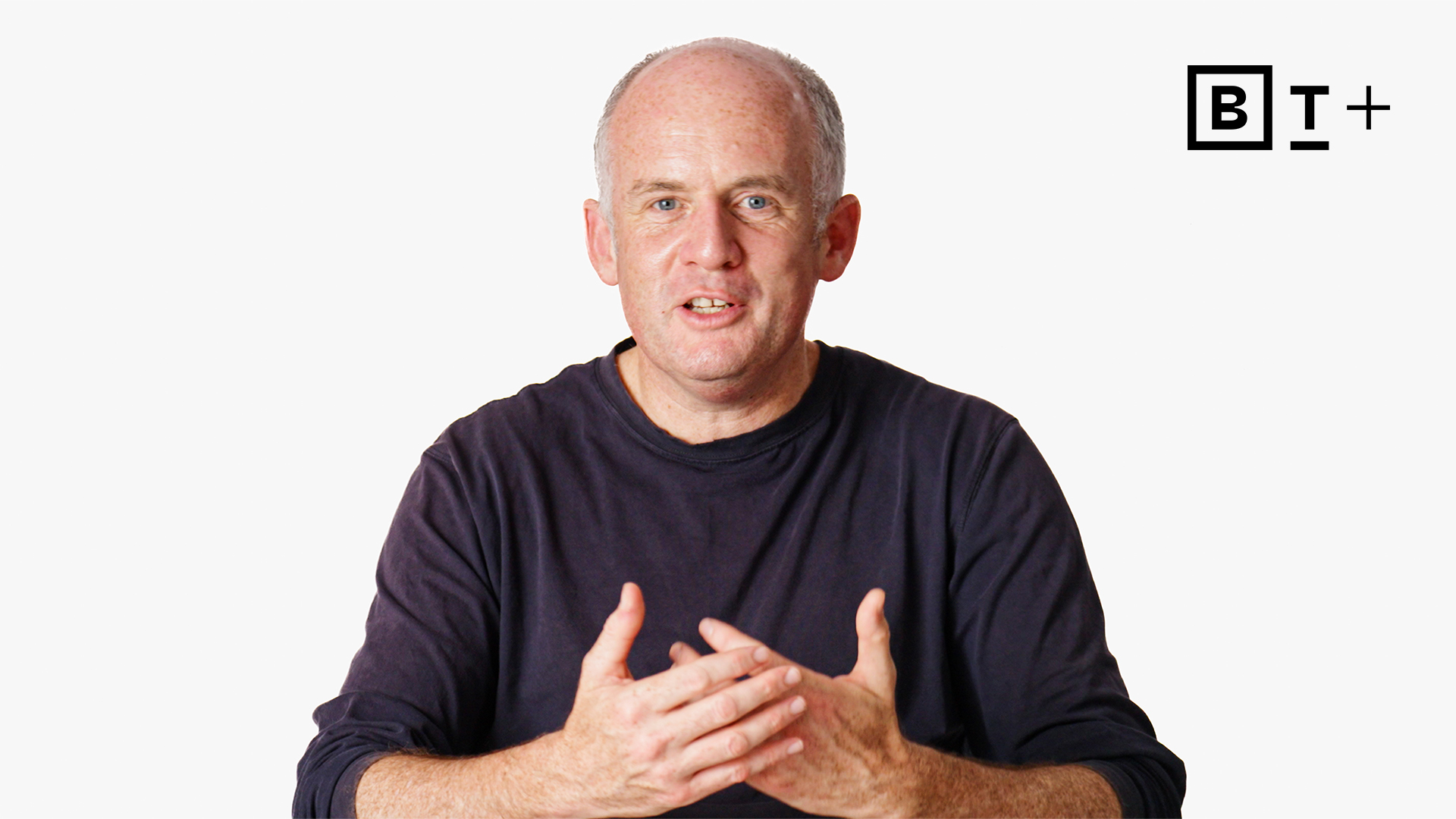 A middle-aged man with short gray hair and a dark shirt speaks with hands raised; a "B T +" logo appears in the top right corner on a white background.