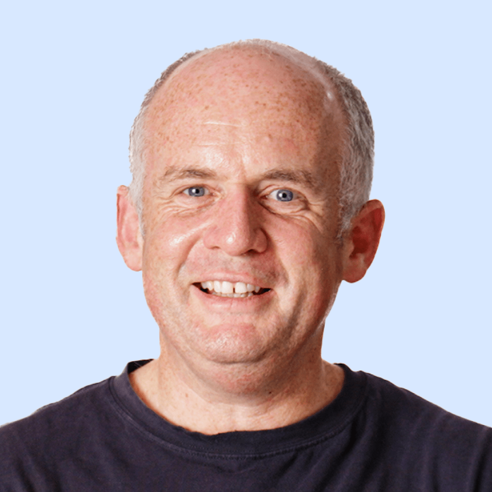 A middle-aged man with short gray hair and a receding hairline is smiling, wearing a dark t-shirt against a light blue background.