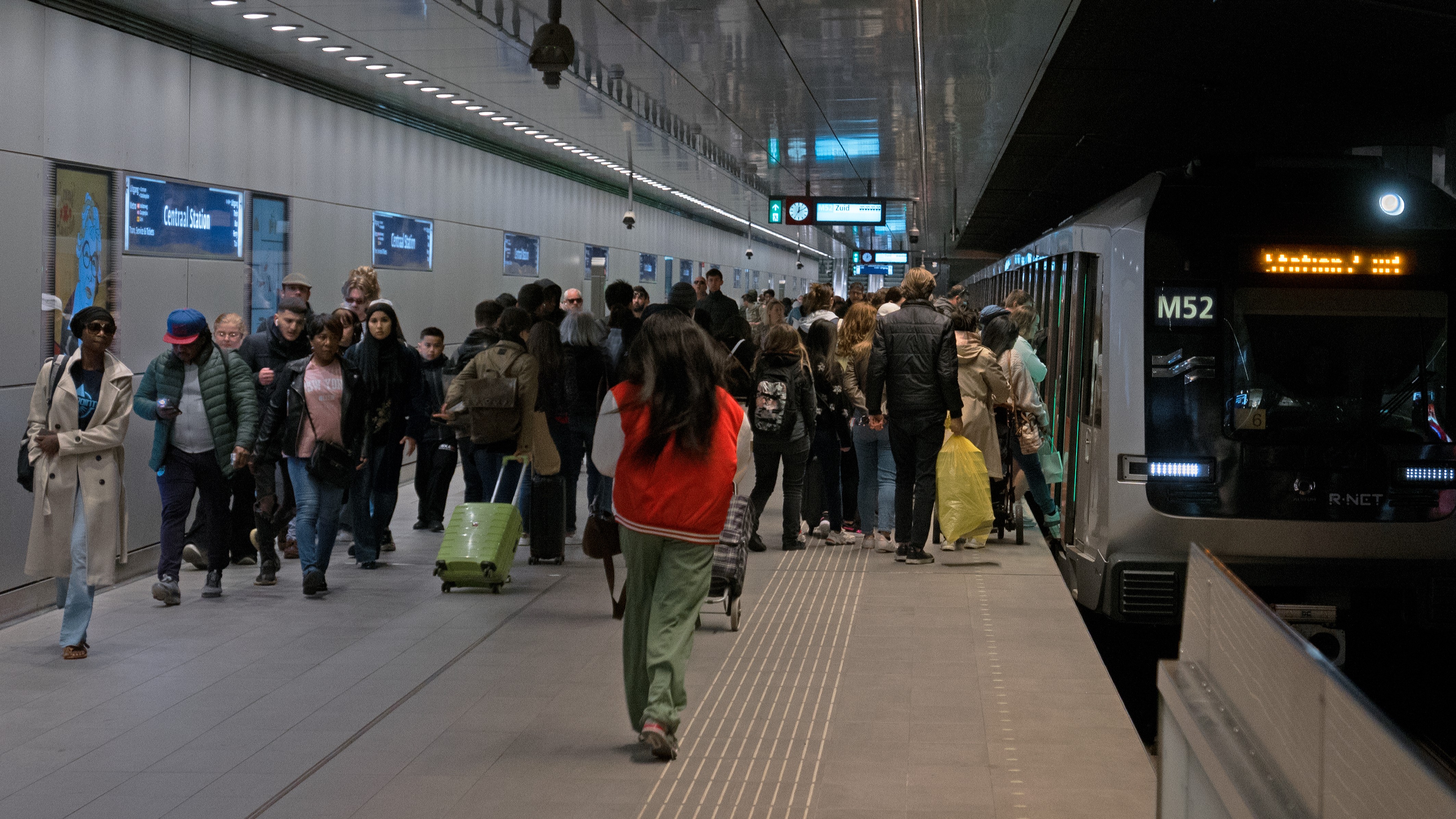 A crowd of people stands and walks on a subway platform as passengers board and exit a train labeled M52.