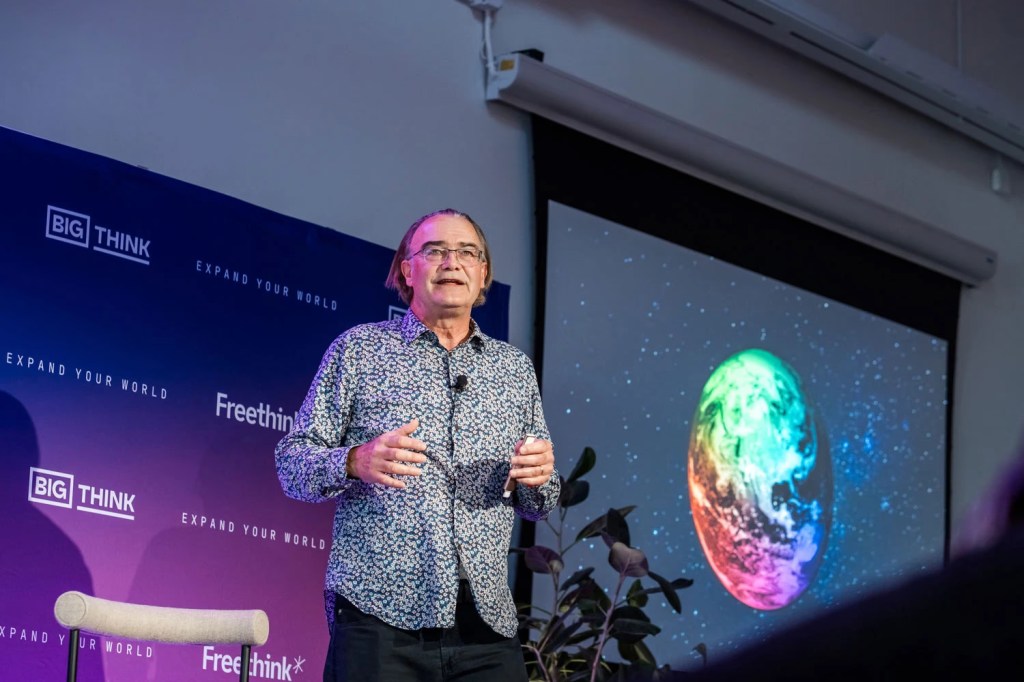 A man stands and speaks on stage next to a projected image of a colorful Earth, with Big Think and Freethink banners behind him.