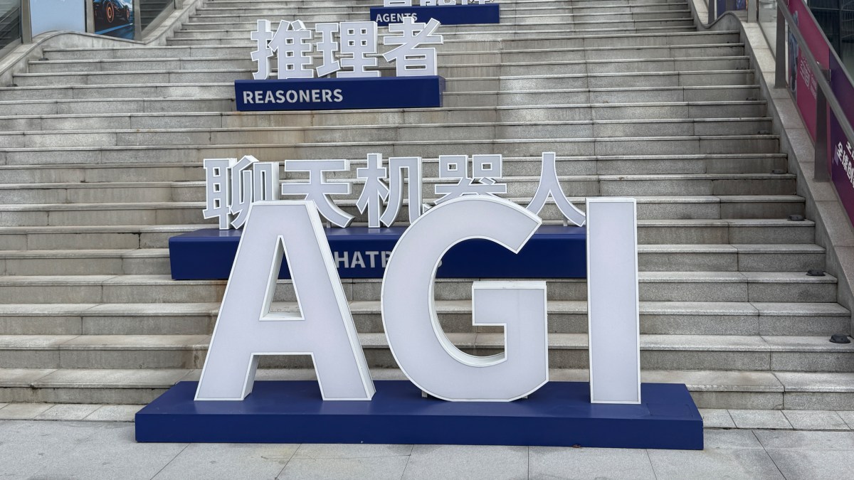 Large white letters spelling "AGI" are displayed on a platform in front of steps, with additional bilingual signs reading "REASONERS" and "CHATBOTS" in English and Chinese.