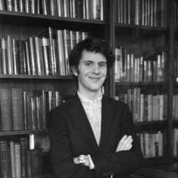 A young man in a suit jacket stands with arms crossed, smiling in front of bookshelves filled with books.