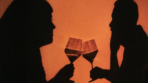 Silhouettes of two people facing each other, clinking wine glasses against an orange background, sharing a lighthearted, possibly drunk moment.