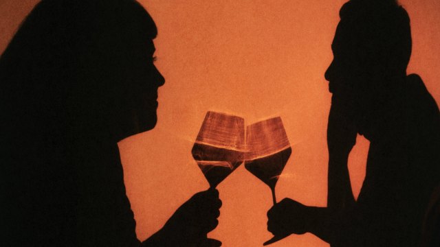 Silhouettes of two people facing each other, clinking wine glasses against an orange background, sharing a lighthearted, possibly drunk moment.