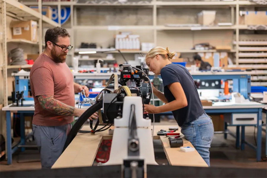 Two people work together on machinery in a workshop, surrounded by tools, equipment, and shelves filled with various supplies.