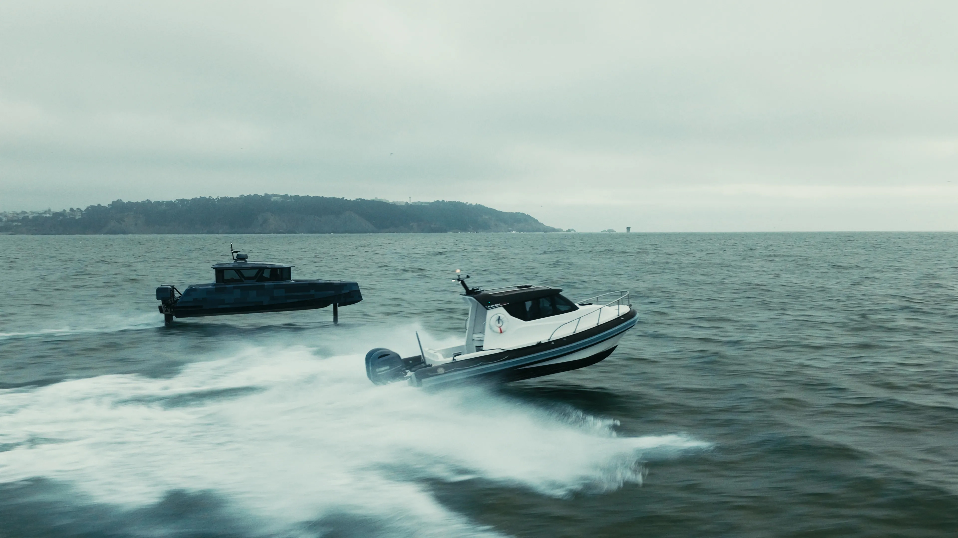 Two motorboats speed across the water under an overcast sky, with a hilly coastline visible in the distance.