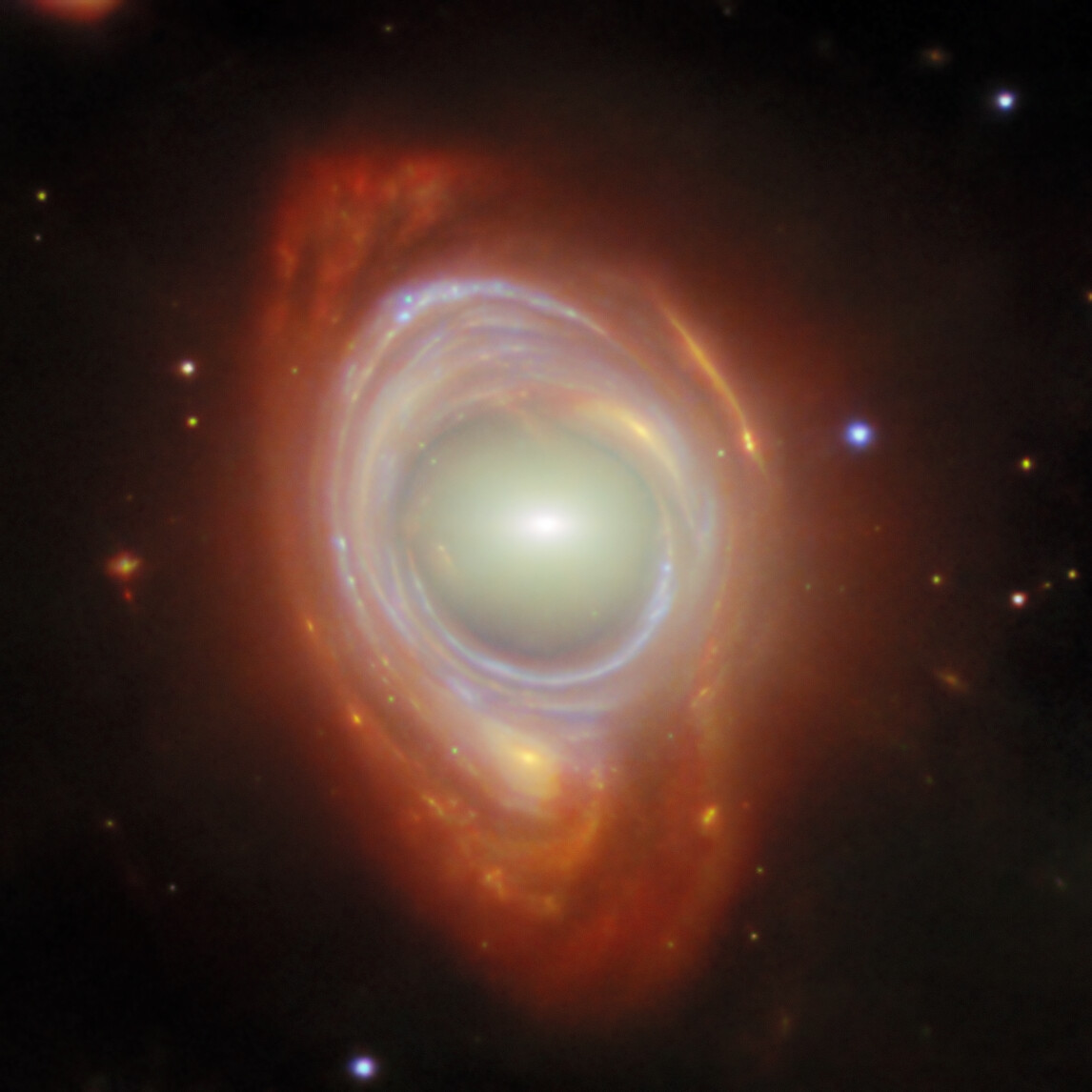 A galaxy with a bright central core, surrounded by glowing reddish-orange rings and a faint outer halo, is captured in the COSMOS-Web JWST survey—its light subtly distorted by a gravitational lens amid scattered stars.