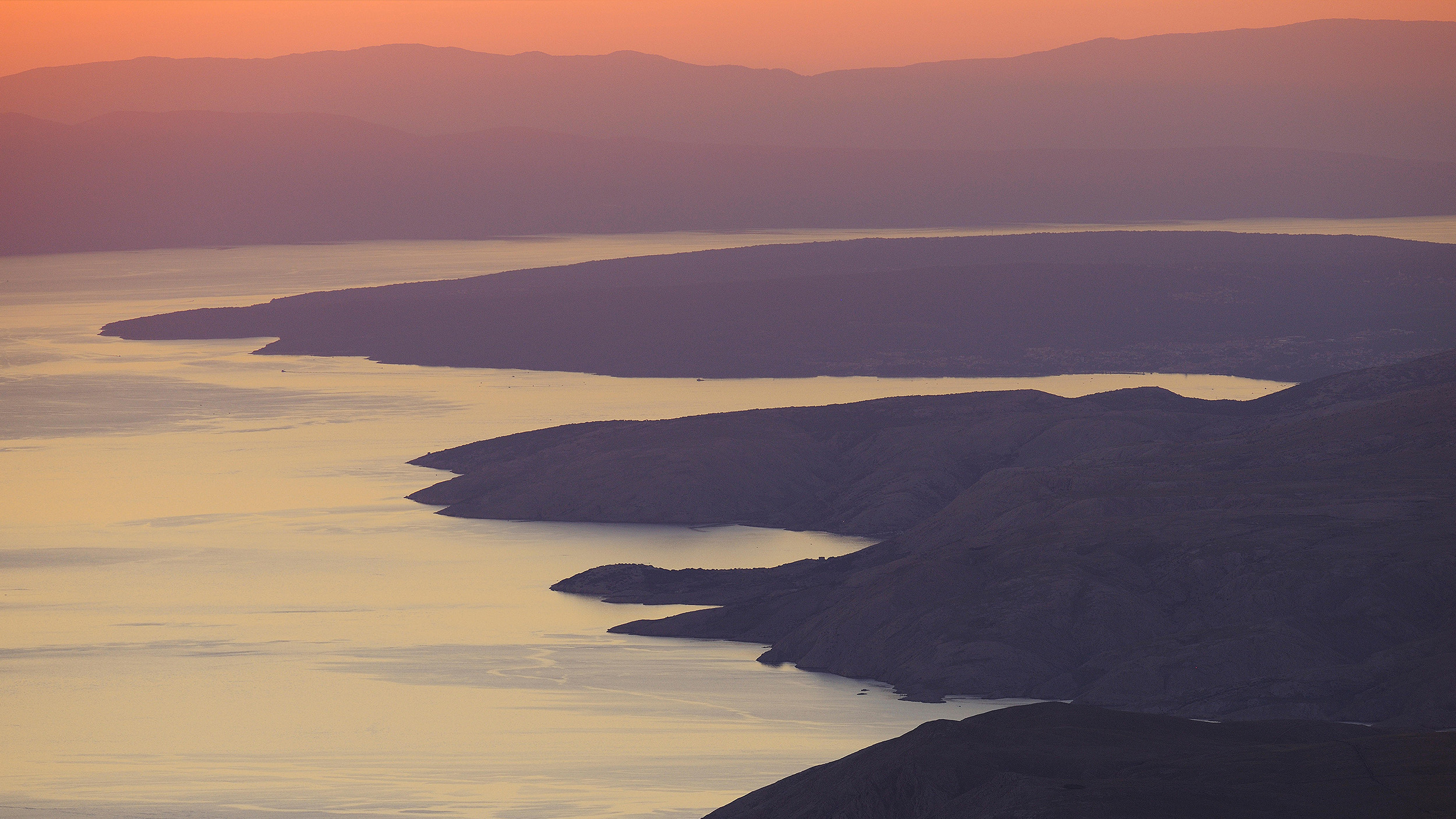 A coastal landscape with rugged cliffs shaped by seaflooding and calm water at sunset, with mountains in the background and soft orange and purple hues in the sky.