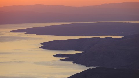A coastal landscape with rugged cliffs shaped by seaflooding and calm water at sunset, with mountains in the background and soft orange and purple hues in the sky.