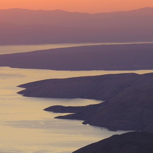 A coastal landscape with rugged cliffs shaped by seaflooding and calm water at sunset, with mountains in the background and soft orange and purple hues in the sky.