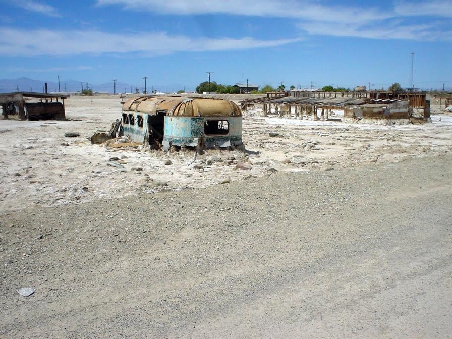 A rusted, abandoned trailer sits on a dry, barren landscape with other dilapidated structures in the background under a blue sky.