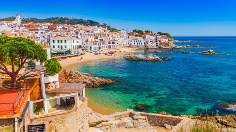 Coastal Mediterranean village with white buildings, a sandy beach, colorful boats, and clear turquoise sea under a bright blue sky.