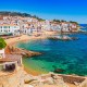 Coastal Mediterranean village with white buildings, a sandy beach, colorful boats, and clear turquoise sea under a bright blue sky.