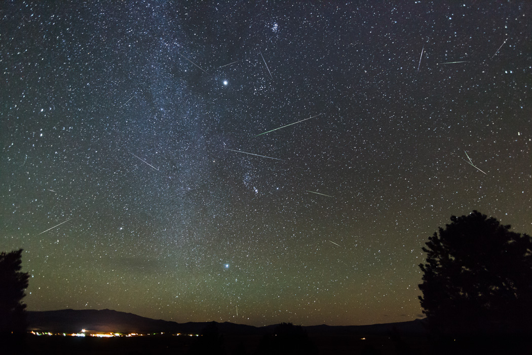 Night sky adorned with countless stars and shooting stars; meteor streaks illuminate the darkness, while the faint silhouette of trees and distant town lights frame the horizon—perhaps a glimpse of Halley's Comet graces the view.