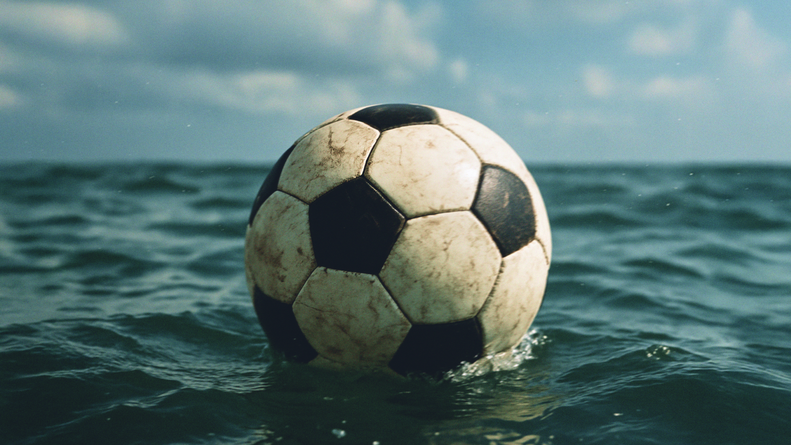 A black and white soccer ball floats on the surface of the ocean under a partly cloudy sky.