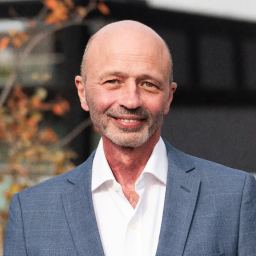 A bald man in a blue suit and white shirt stands outdoors in Silicon Oasis, smiling, with autumn leaves and a blurred building in the background.