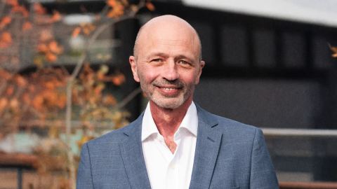 A bald man in a blue suit and white shirt stands outdoors in Silicon Oasis, smiling, with autumn leaves and a blurred building in the background.