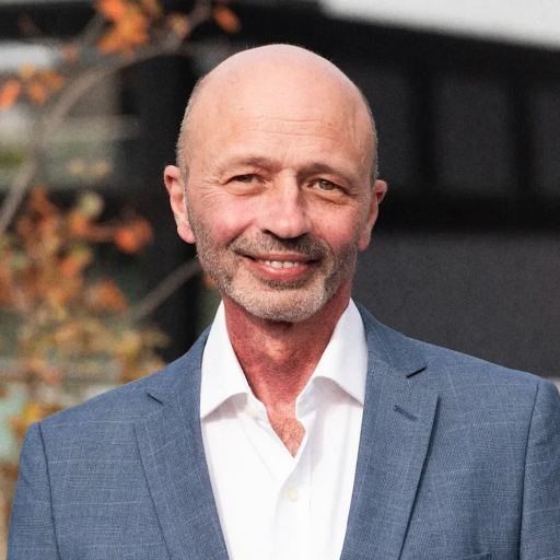 A bald man in a blue suit and white shirt stands outdoors in Silicon Oasis, smiling, with autumn leaves and a blurred building in the background.